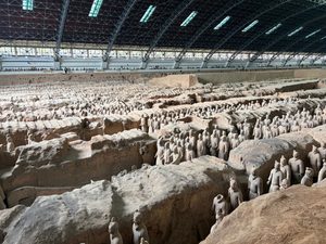 Rows of life-sized Terracotta Army warriors standing in excavated pits beneath the large covered archaeological museum hall in Xi’an, China, with visitors observing from elevated walkways.