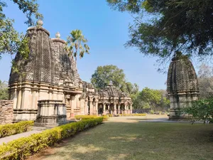 Historic stone temple complex in India featuring intricately carved towers, pillared structures, landscaped gardens, palm trees, and surrounding greenery beneath a clear blue sky.
