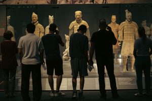 Visitors standing in front of a glass museum display featuring life-sized Terracotta Army warrior statues, including armored soldiers and additional ancient figures illuminated by exhibition lighting.