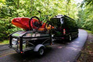 🚐✨ Comfort & Convenience: 50 RV Upgrades For Stress-Free Travel📝 5 Camper towing a trailer with a kayak and bicycle secured on top, driving through a scenic outdoor area.