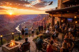 Scenic Grand Canyon patio restaurant overlooking the Colorado River at sunset with people dining outdoors, guests standing at the railing viewing canyon scenery, a fire pit, glowing lanterns, and an eagle flying overhead.