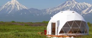 🎄✨ Holiday Glamping Must-Haves: 50 Products For Festive Outdoor Getaways 1 White glamping dome on a platform in a field with large snow-covered mountains in the background.
