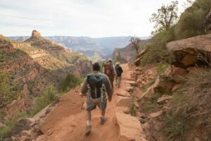 Three hikers with backpacks walk along a ridge at the top of a valley, surrounded by rolling hills and sweeping vistas under bright sunlight.