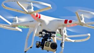 White drone with red markings and green lights flying in a clear blue sky, equipped with a camera.