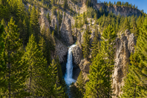 Tall waterfall cascading through a rugged forested canyon surrounded by evergreen trees and rocky cliffs beneath a bright blue sky.