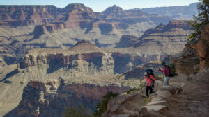 Two hikers with backpacks walking down a trail into the Grand Canyon surrounded by steep cliffs and rocky terrain.