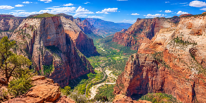 Wide panoramic view of Zion National Park featuring towering red sandstone cliffs, a lush green canyon valley, and rugged desert scenery under a bright blue sky.