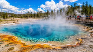Vibrant Yellowstone geothermal hot spring with crystal-clear blue water, rising steam, colorful mineral edges, and visitors standing on a wooden boardwalk beneath a bright partly cloudy sky.