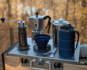 A stylish outdoor setup with coffee-making gear — French press, mugs, and accessories — displayed on a camping or glamping table.