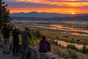 Visitors using binoculars and a camera to watch bison across Hayden Valley at dusk, with winding waterways, mountains, and a colorful sunset.