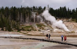 Visitors walking along a wooden walkway with steam rising, a hill, and forest in the background.