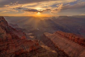 Sunlight breaking through clouds over a steep canyon with layered cliffs and a deep valley, highlighting the textures of the rock formations.