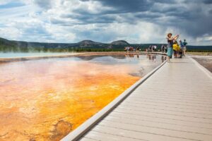 “Visitors walking on a wooden boardwalk at eye level near a Yellowstone geyser, with blue water and many people around.