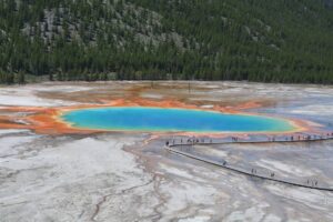 Geyser in Yellowstone with visitors walking on a boardwalk, showing blue water and gray mineral deposits.