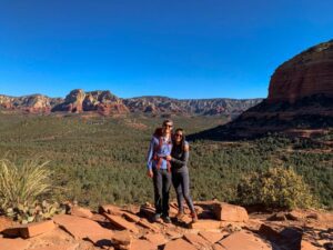 Two people standing in a desert landscape surrounded by small shrubs with mountains in the distance.