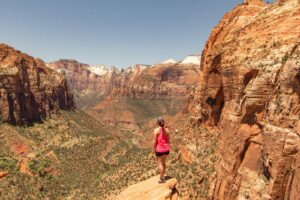 Exploring Zion National Park: Utah’s Majestic Canyon Wonderland 5 A woman stands on a rocky ledge overlooking a vast canyon below, wearing a backpack and gazing into the dramatic landscape filled with cliffs and sunlight.