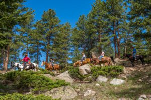 Estes Park – Gateway to The Rockies and Outdoor Adventure 2 People riding horses through a forested mountain landscape with trees and peaks in the background.