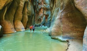 Exploring Zion National Park: Utah’s Majestic Canyon Wonderland 3 A group of hikers walks through the Narrows in Zion National Park, wading in shallow river water between towering red canyon walls glowing in soft light.