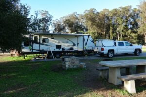 A truck and trailer parked in a scenic South America RV site with a picnic table and fire pit, surrounded by lush trees, ready for a camping adventure.
