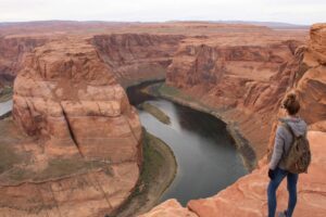 Woman standing on a rocky ledge overlooking a deep canyon with a river flowing through the valley below under a clear sky.