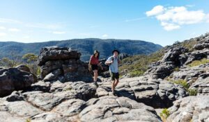Two hikers with backpacks trekking along large rocky terrain surrounded by mountains.