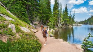 Two hikers with backpacks walking along Emerald Lake Trail in Rocky Mountain National Park, next to a lake with forest and mountains nearby.