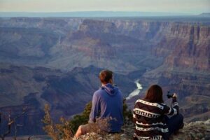 Two people sitting at the top of a canyon, looking down at a flowing river winding through the steep canyon walls under clear skies.