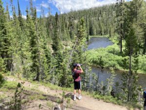 Person standing on a trail above a river, looking through binoculars at the surrounding forest and mountains.