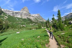 Person hiking on a trail with a backpack, surrounded by forest and mountain scenery.