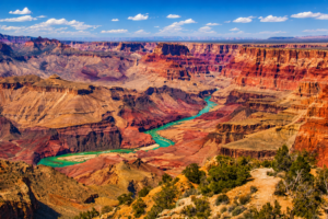 Panoramic view of the Grand Canyon South Rim featuring towering red rock cliffs, colorful layered canyon walls, and the winding Colorado River stretching through the canyon beneath a bright blue sky.