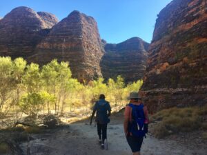 Two hikers with backpacks walking along a trail near large rock formations.