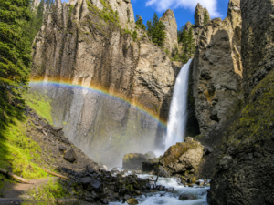Tower Fall – Yellowstone’s Classic Waterfall 5 Tower Fall cascading through rugged volcanic cliffs with a colorful rainbow, evergreen trees, and rocky stream scenery in Yellowstone National Park.