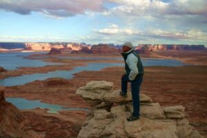 🌊 Lake Powell (Page, Arizona) Waterfront RV Camping 3 Man standing on a rocky outcrop overlooking flatlands, a lake, and mountains in the distance under a clear sky.
