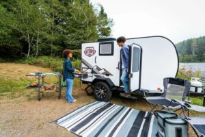 🚐✨ Comfort & Convenience: 50 RV Upgrades For Stress-Free Travel📝 3 Two people sitting outside a small camper with a rug and portable generator at a campsite.