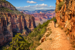Solo hiker walking along a narrow canyon trail surrounded by towering red rock cliffs and breathtaking panoramic views deep inside Grand Canyon National Park beneath a bright blue sky.