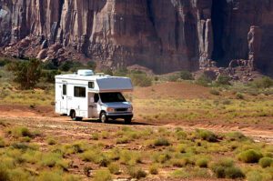 Camper driving across flat desert land flanked by steep sandstone cliffs.