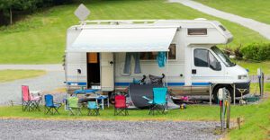 White camper with blue stripe parked outdoors, surrounded by multiple folding chairs in front, ready for outdoor relaxation.