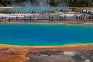 Grand Prismatic Spring in Yellowstone National Park with brilliant blue geothermal water, orange mineral deposits, rising steam, and visitors walking along surrounding boardwalks.