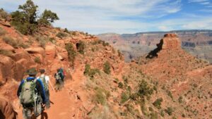 Four people with backpacks walking along a trail at the top of a canyon, surrounded by expansive cliffs and rocky terrain.