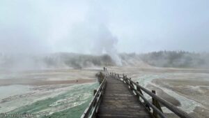 Visitors walking on a bridge walkway with steam rising from a Yellowstone geyser below.