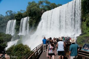 Several people standing on a walkway with railings at the base of waterfalls surrounded by lush rainforest vegetation.