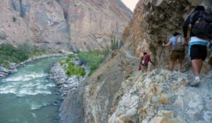 Several hikers wearing backpacks walking along a trail near a creek, surrounded by lush vegetation and mountains.