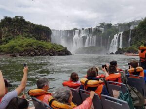 Several people wearing life jackets rafting on a river near cascading waterfalls surrounded by lush vegetation.