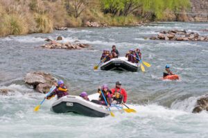 Several people wearing helmets rafting downstream through rapids on a river surrounded by lush scenery.