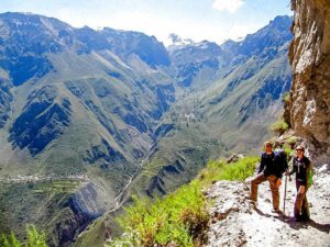 Two hikers wearing backpacks walking on a mountain trail high above a deep valley with steep cliffs and distant peaks.