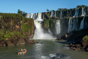 Boat cruising on a river through the lush rainforest with Iguazú Falls visible in the background, surrounded by tropical vegetation.