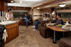 Family relaxing inside a cozy RV camper with comfortable seating, dining area, kitchen space, warm lighting, and scenic forest views through the windows during a peaceful camping trip.