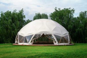 White glamping dome set in a lush green field with a forest backdrop, showcasing a serene outdoor getaway.