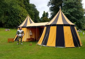 🌞🌈🏔️🌿Unique & Novelty Tents: 30 Medieval, Tipi & Yurts for Glamping & Festivals🎪🔥 3 black and yellow Medieval tent wioth person in armored suit on left with trees in the background