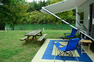 RV camper parked with a rug, chairs, and picnic table in front, beside a flowing river with trees